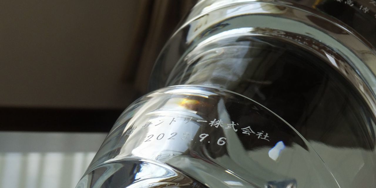 バカラの名入れ花瓶一覧で使用する写真。バカラのスパイラルベースの側面に、『贈 サントリー株式会社 日付』を名入れ彫刻したアップ写真。螺旋状の美しいフォルムを持つ花瓶に、企業からの贈答メッセージが繊細に刻まれ、企業の品格と感謝の気持ちを伝える、洗練された記念品としての価値を際立たせています。
