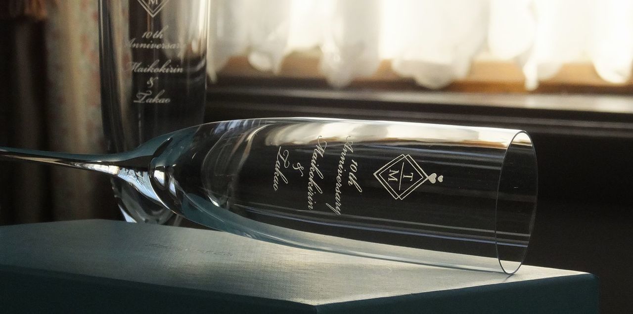 ティファニー（Tiffany & Co.）のホームエッセンシャル シャンパングラスのペアセットのクローズアップ写真。細くエレガントなステムを持つグラスの側面中央に、結婚10周年記念として**『2人のイニシャル』、『ハートのデザイン』、『10th Anniversary』、そして『2人の名前』が名入れ彫刻されているのが鮮明に確認できる。ブランドの格調高さと、10年の愛の軌跡を祝うロマンチックなデザインが融合し、永遠の誓いを象徴する特別な記念品。