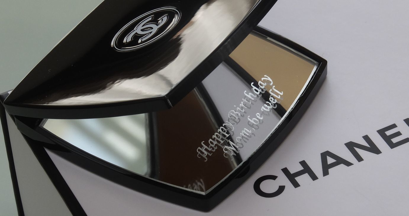 シャネル（CHANEL）のコンパクトミラーのクローズアップ写真。誕生日プレゼントとして贈られた、黒く象徴的なケースを開いた鏡面、あるいは裏側に、『Happy Birthday Mom, be well』という母親への誕生日と健康を願うメッセージが繊細に名入れ彫刻されているのが鮮明に確認できる。ブランドの高級感と、母への深い愛情と感謝を伝える心温まるメッセージが込められたエレガントでパーソナルなカスタムギフト。