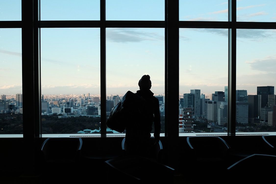 新しい人生の始まりを象徴するシーン。高層ビルの窓から都市のビル群を眺める男性が、大きなボストンバッグを手にしている。未来に向けての決意と期待感が感じられる瞬間