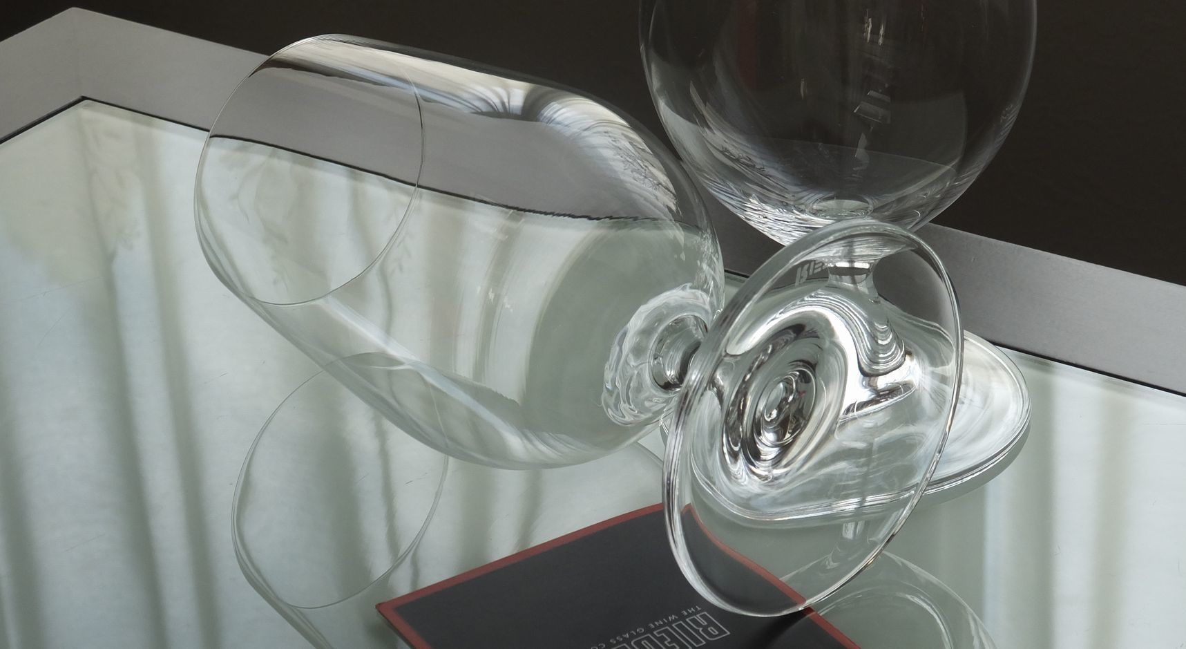 テーブルの上に横向き（倒した状態）で置かれた、リーデル（RIEDEL）の『オヴァチュア ビアグラス』の写真。ボウルの膨らみや繊細なステム（脚）、最高級クリスタルならではのなめらかな曲線の美しさと透き通る透明感がディテールまで際立つイメージ。