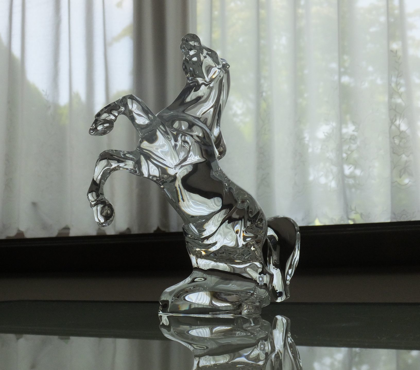 洗練されたテーブルの上に置かれた、バカラ（Baccarat）のクリスタル製オブジェ『いななく馬』の写真。前足を高く上げ、今にも動き出しそうなほど生命力と躍動感にあふれる美しいシルエット。最高級クリスタルガラスならではの圧倒的な透明感と滑らかなカットが光を均一に反射し、空間に極上の輝きと品格をもたらす。栄誉や成功、躍進を象徴する格調高いフィギュリンとして、特別な記念日や法人向けの最高峰のギフトにふさわしいラグジュアリーなイメージ。