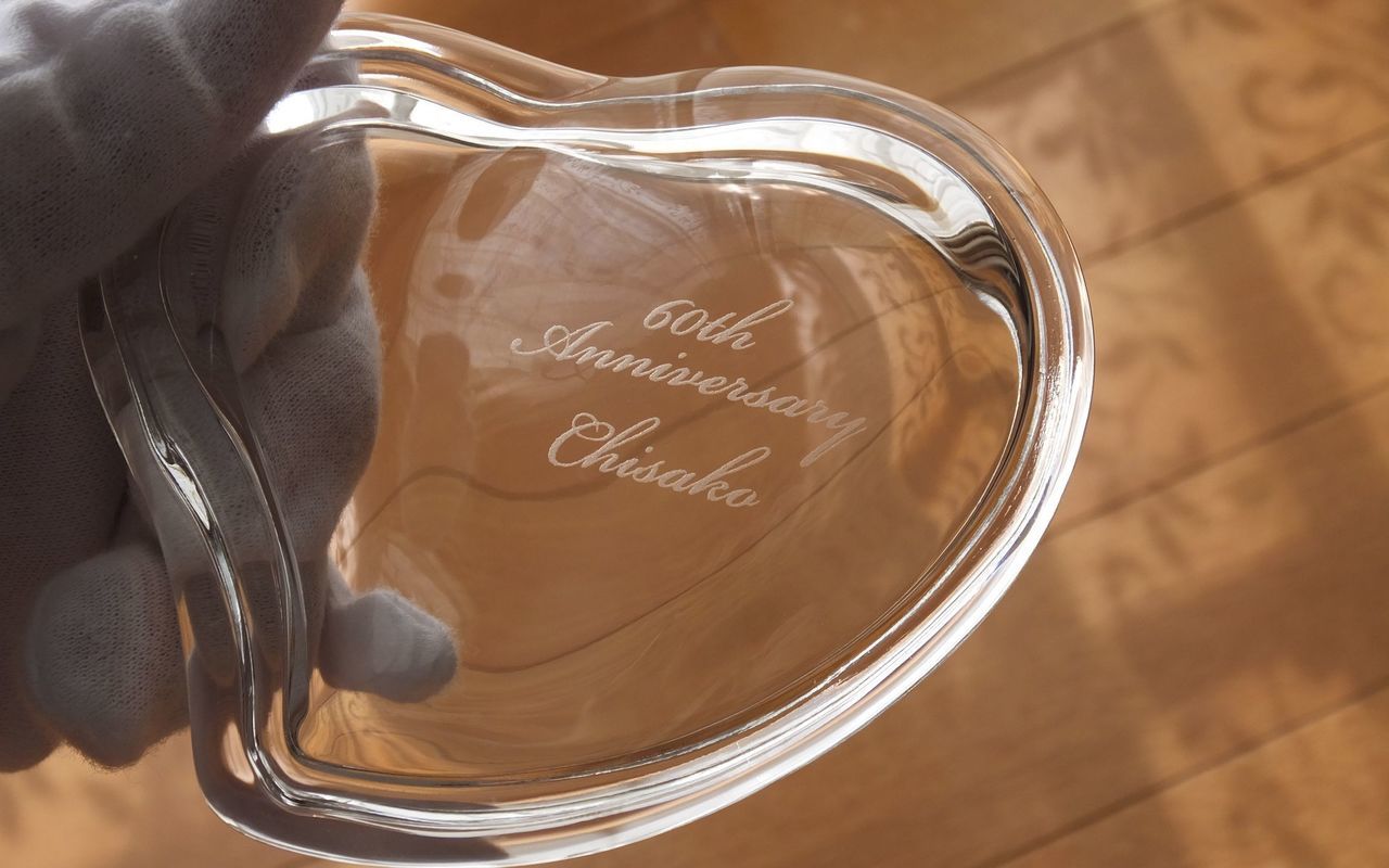 ティファニー（Tiffany & Co.）の『クリスタル ハート ボックス』の蓋部分に、『60th Anniversary Chisako』の還暦祝いメッセージとお名前が精緻に名入れ彫刻されたお祝いの写真。愛らしく可憐なハート型のフォルムとティファニーならではの上質な透明感あふれるクリスタルガラスに、上品でエレガントな刻印が美しく浮かび上がり、人生の節目である還暦（60歳）の特別な祝福と心からの敬意を格調高く伝えるメモリアルギフトイメージ。」