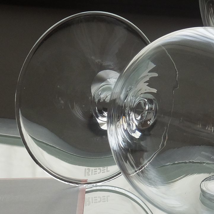 テーブルの上に置かれたリーデル（RIEDEL）の『オヴァチュア ビアグラス』の底面に、ブランドの証である『RIEDEL』ロゴ刻印が大きく拡大された写真。最高級クリスタルガラスならではの繊細で美しい透明感とともに、本物の品質と伝統を証明する上品なロゴが際立ち、特別なお祝いや記念品などの贈り物にふさわしい信頼感あふれるイメージ。
