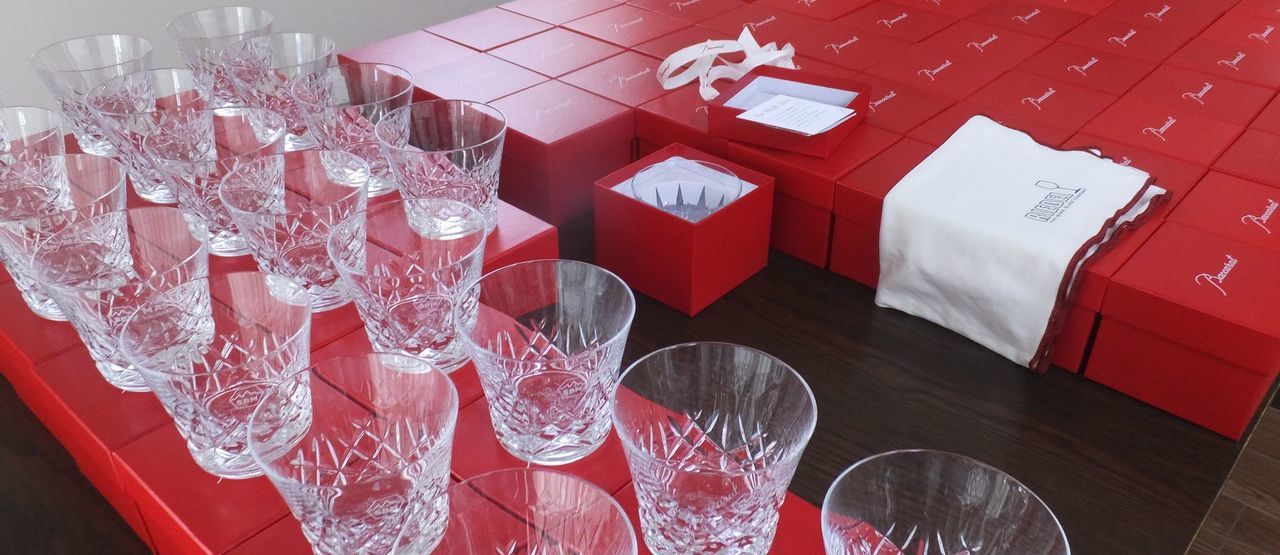 バカラのアリア（Aria）タンブラー2025が、品質管理または検品作業のために、真紅のオリジナルボックスの上に50個以上、整然と規則正しく並べて置かれている集合写真。しずくを思わせる繊細なカットが施されたグラスが、均一な配置と光の反射により壮観な輝きを放っている。クリスタル製品の最高品質を保つための厳格なチェックを象徴する情景であり、大量注文の完璧な仕上がりを伝える。