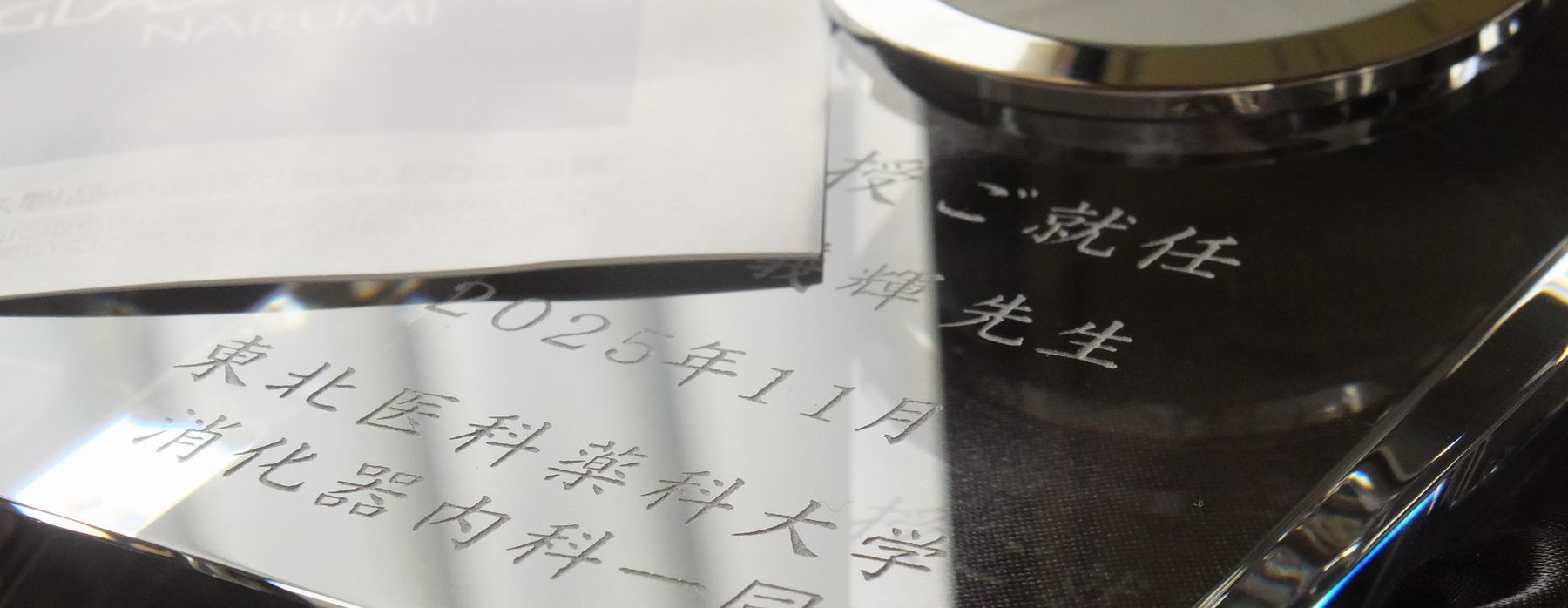 グラスワークスナルミのエンバシークロックに、『教授ご就任』の祝辞、先生のお名前、『2025年11月 東北医科薬科大学 消化器内科一同』と名入れ彫刻を施した写真。透明度の高い光学クリスタルに、医局員一同からの敬意と祝意が刻まれた、就任祝いに相応しい格調高い記念品のイメージ。