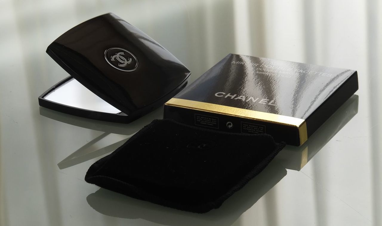 シャネルのコンパクトミラーの一式紹介写真：箱、ミラー本体、そしてミラーを収納するベロア風の袋が、テーブルの上に美しく配置された写真。ギフトとしての完全なセットと、ブランドの細部へのこだわりを伝えます。