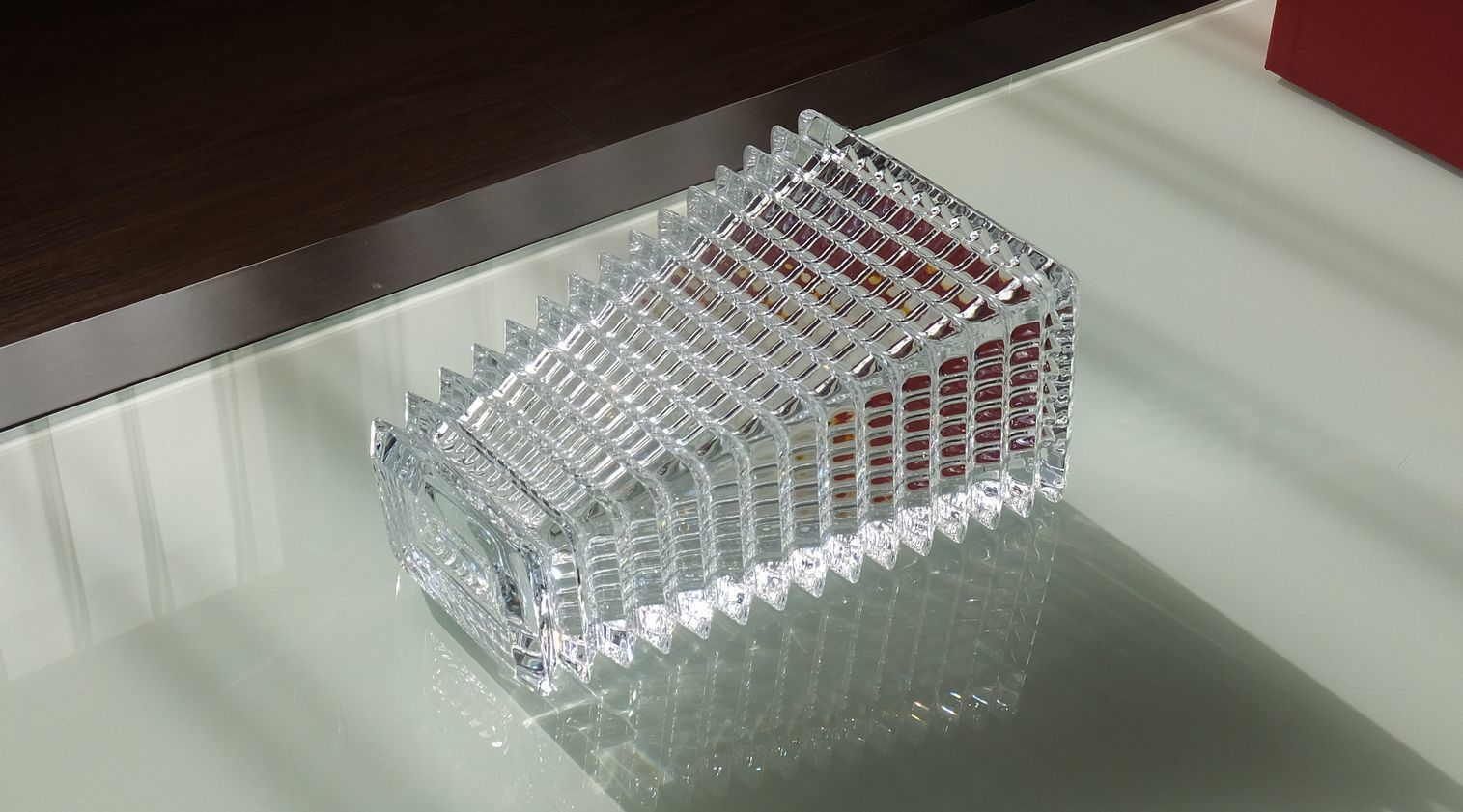 バカラの『アイ ベース スクウェア（角型）』が、モダンなガラステーブルの上に横向きに倒して置かれている様子の写真。レンズのカットをイメージした内外の凹凸が、光の屈折と影を複雑に作り出し、横倒しにすることでその視覚効果が強調されている。特に、透明なクリスタルが透明なガラス面と接することで、ベースの幾何学的なカットの美しさと、横向きならではのダイナミックな光の反射が最大限に引き出され、オブジェのような芸術的な存在感を放っている。