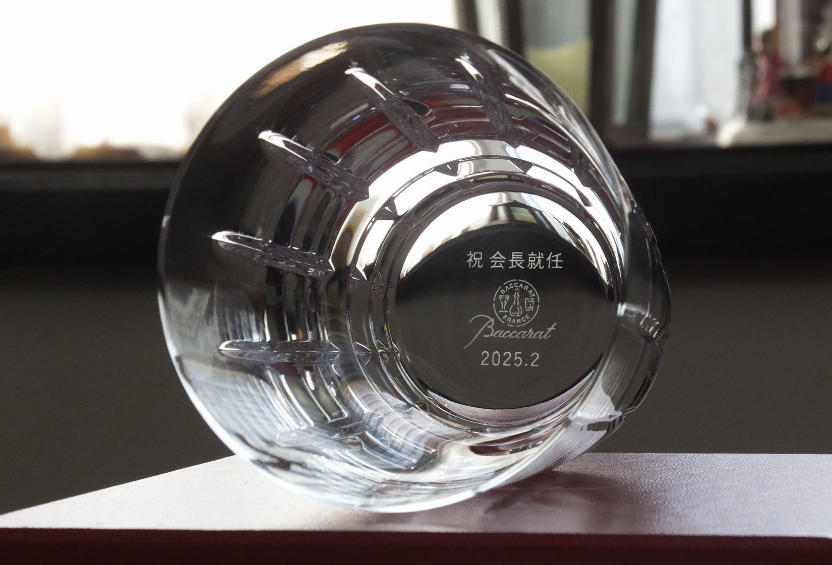 バカラ（Baccarat）の『アルルカン タンブラー』の底面に、『祝 会長就任』の文字と『2025.2』の記念日付が精緻に名入れ彫刻された写真。道化師の衣装をモチーフにした幾何学的なカットワークが光を華やかに反射する最高級クリスタルグラスの底から、新たな門出と栄誉を讃える刻印が密やかに美しく輝き、役員就任や会長就任祝い、特別な企業表彰に贈る最高峰の法人記念品にふさわしい格調高いイメージ。