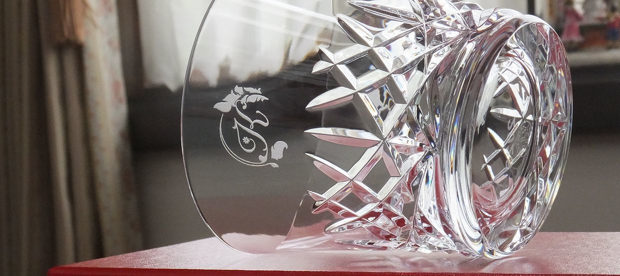 バカラのアリア（Aria）タンブラーのクリスタルグラスのクローズアップ写真。グラスの側面中央に、誕生日プレゼントとして『イニシャルデザイン K』の文字が繊細に名入れ彫刻されているのが鮮明に確認できる。しずくを思わせる繊細なカットが特徴のタンブラーの透明度の高い面に、パーソナルなイニシャルが際立ち、世界最高峰の輝きとともに特別な誕生日を祝う洗練されたカスタムギフト。