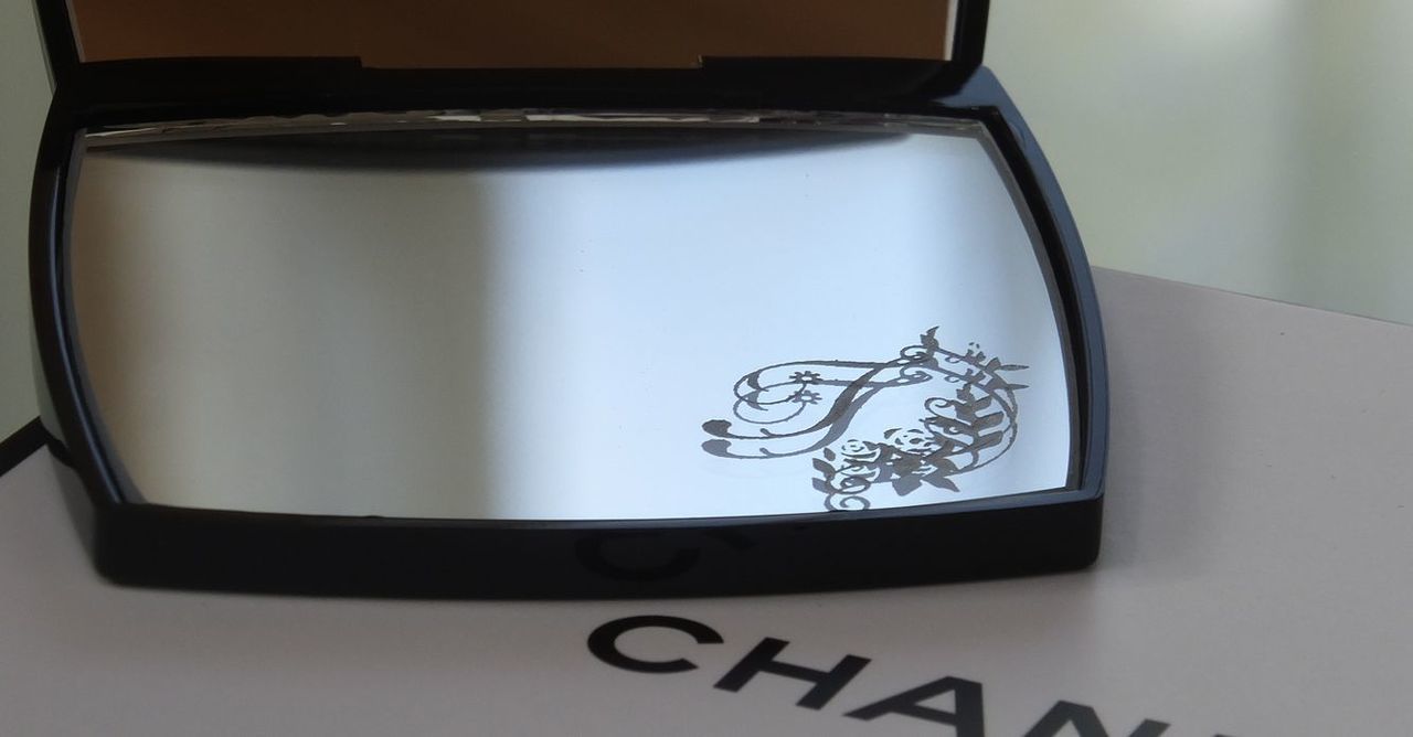 シャネルのコンパクトミラーの下部ミラーに、エレガントなバラの装飾とイニシャル『T』を名入れ彫刻した写真。華やかで繊細なデザインが高級感を醸し出す、特別なギフト仕様のイメージ。