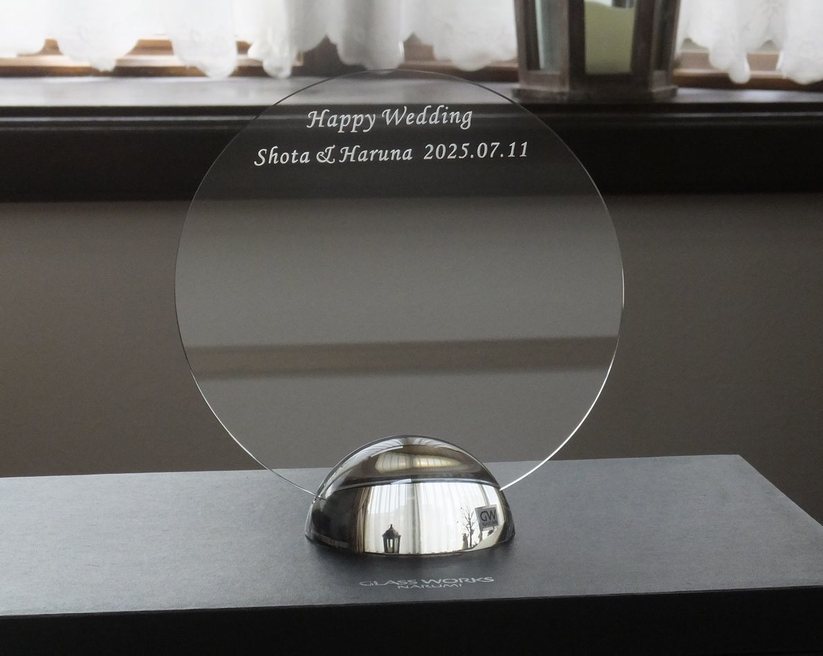 グラスワークスナルミのクリスタル製のドームフォトフレーム（写真立て）が、テーブルの上に置かれている写真。緩やかなアーチを描くクリスタルの下部には、結婚祝いのメッセージとして『Happy Wedding Shota & Haruna 2025.07.11』という新郎新婦の名前と記念日が名入れ彫刻されているのが確認できる。透明度の高いフレームが周囲の光を集め、結婚という特別な節目の幸せな思い出をエレガントに彩る**、パーソナルな記念品の情景。