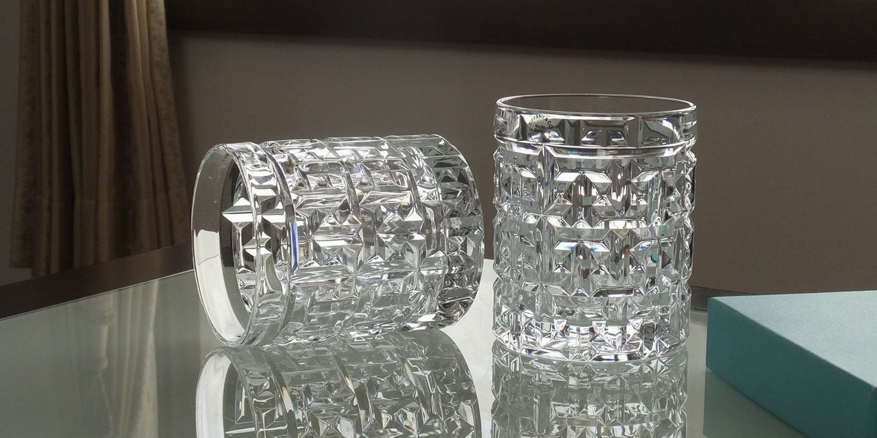 ティファニー（Tiffany & Co.）のクリスタルグラス、T トゥルー ダブル オールドファッションのペアグラス（2個）が、テーブルの上に並べて置かれている写真。グラス全体に垂直に施された細かなラインカットと、グラスの底面近くに象徴的に刻まれた『T』のモチーフが特徴的。透明度の高いクリスタルが光をシャープに反射させ、モダンで洗練された重厚感を演出している。結婚祝いや特別な記念日の贈り物にふさわしい、エレガントなペアの情景。