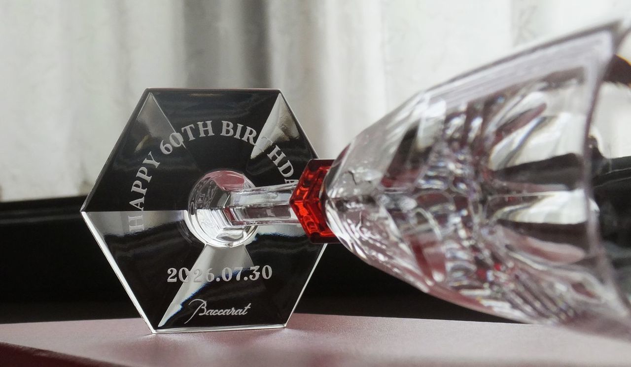 バカラ（Baccarat）の『アルクール イヴ シャンパングラス』の底面に、『Happy 60th Birthday』の祝い文字と記念日付が精緻に名入れ彫刻された還暦祝いの写真。アイコニックなフラットカットの重厚感にフェミニンでエレガントな繊細さを加えた最高級フルートグラスのフット（底面）から、人生の大きな節目を祝う言葉が美しく浮かび上がり、60歳の特別なバースデーや赤を象徴する長寿の記念にふさわしい最高峰のラグジュアリーギフトイメージ。
