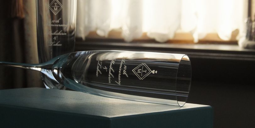 ティファニー（Tiffany & Co.）の最高級クリスタル製シャンパングラスの側面に、優美に洗練されたお二人のイニシャルデザイン、『10th Anniversary』の記念文字、そして流麗なスクリプト体（英文流書体）でお二人のお名前が精緻に名入れ彫刻された結婚10周年記念（錫婚式・アルミ婚式）ギフトの写真。エレガントなフォルムに透明感あふれる光が煌めき、特別な10周年の節目を祝う心躍る記念品や夫婦の絆を刻むラグジュアリーなパーソナルギフトイメージ。