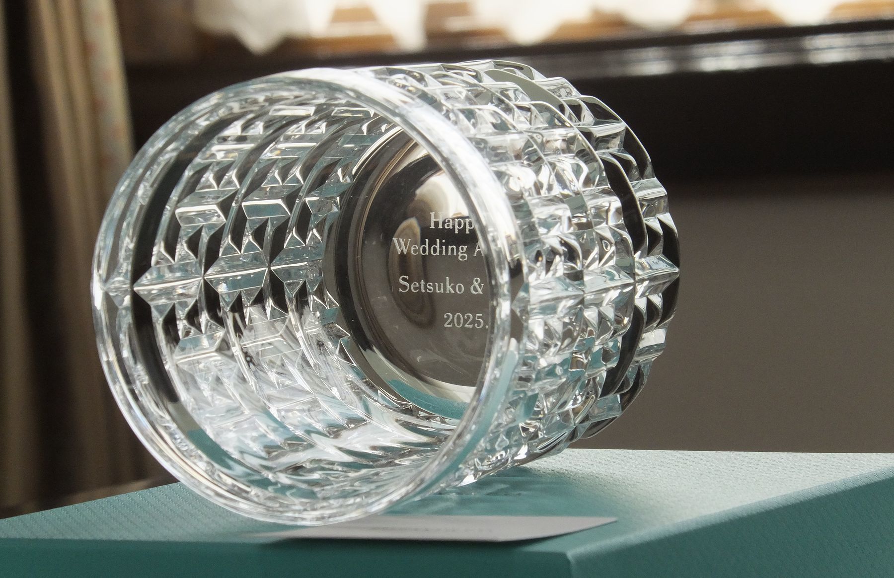 ティファニー（Tiffany & Co.）のTトゥルー ダブルオールドファッションのペアグラスのクローズアップ写真。モダンで構築的なT字モチーフのカットが特徴の重厚なグラスの底面中央に、結婚祝いとして**『Happy Wedding Anniversary』、『二人の名前』、そして『日付』が名入れ彫刻されているのが鮮明に確認できる。ブランドの象徴的なデザインと、底面に秘められた二人の愛の誓いが融合し、新しい生活の始まりを祝うモダンで格調高いギフト。