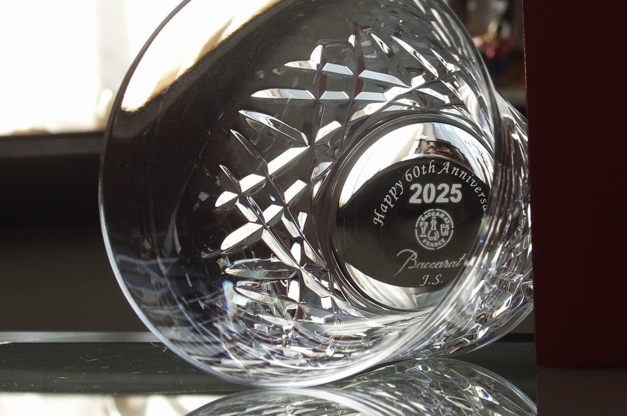 バカラ（Baccarat）の2025年年号刻印入りイヤーグラス『アリア タンブラー』のグラス底面に、『Happy 60th Anniversary J.S』の還暦祝いメッセージとイニシャル（お名前）が精緻に名入れ彫刻されたお祝いの写真。波打つような優美な縦ラインのカットワークの中心部からクリアに浮かび上がる刻印と最高級クリスタルガラスの誇り高き輝きが、人生の大きな節目である還暦（60歳）の祝福とこれからの健やかな未来への祈りを格調高く表現するメモリアルギフトイメージ