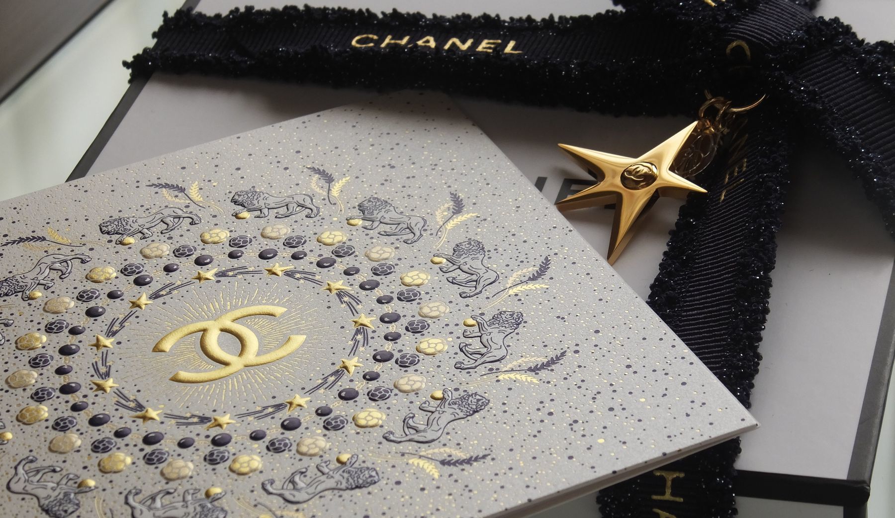 シャネル（CHANEL）のコンパクトミラーに名入れ彫刻を施し、クリスマスシーズンの特別仕様で梱包されたギフトボックスの写真。黒を基調とした高級感のある箱を開けると、鏡の表面または裏側に『[名前]』や『Merry Christmas』といった名入れメッセージが確認できる。箱の外側にはゴールドまたはシルバーのスター（星）モチーフのチャームと、手書き風のメッセージカードが添えられており、ブランドの格調高さと特別な贈り物としての華やかさを伝える情景。