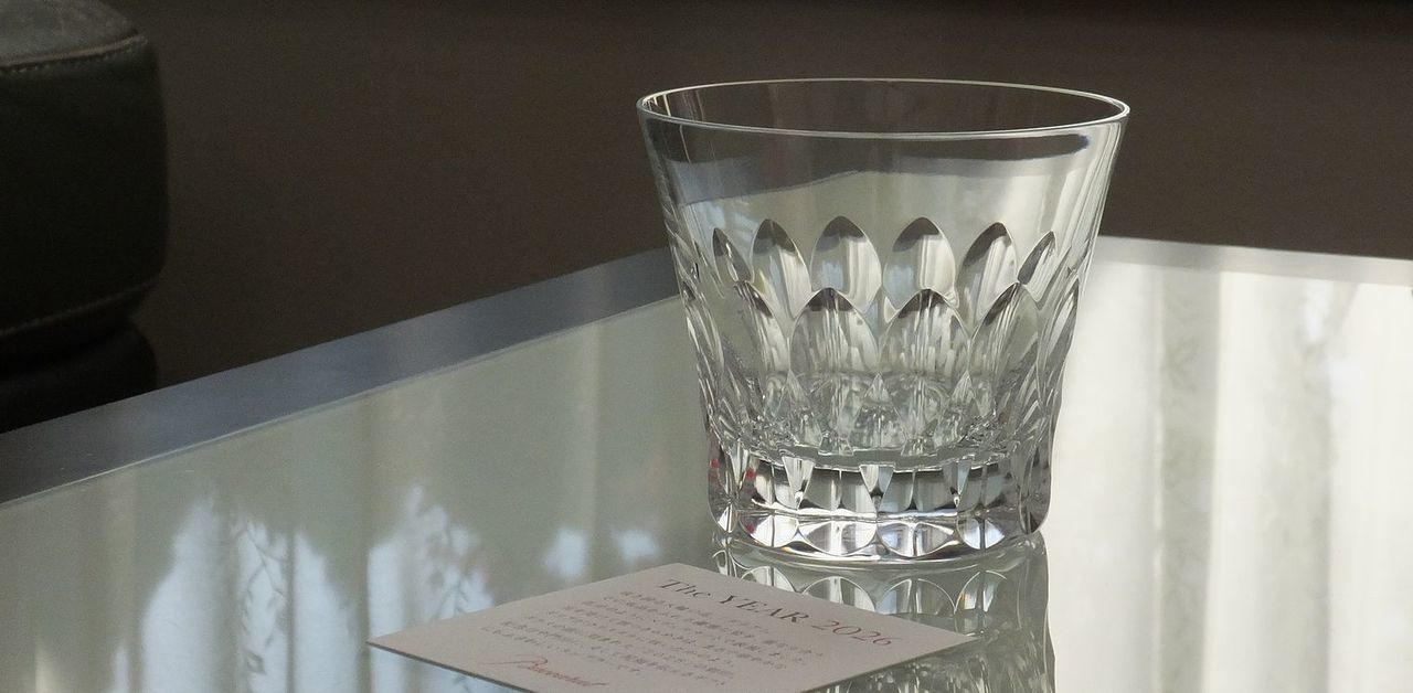 バカラのイヤーグラス、ダリア（Dahlia）タンブラー2026のクリスタルグラスが、テーブルの上に置かれている写真。ダリアの花弁をモチーフとしたダイナミックかつ複雑なV字カットが、室内の光を捉えて多方向に反射させ、グラス全体が眩いばかりの輝きを放っている。重厚な底面と優美なカットが、クリスタルアートとしての存在感を強調し、特別な記念を彩る世界最高峰のタンブラーの情景。