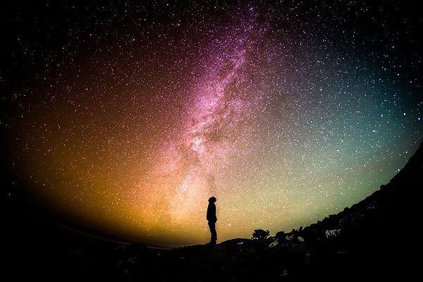 一人の男性が夜空を見上げ、無数の星々が輝き、中央には天の川が広がる美しい星空を眺めるシーン。壮大な宇宙の中で、静けさと広がる神秘的な光景に心を奪われる瞬間