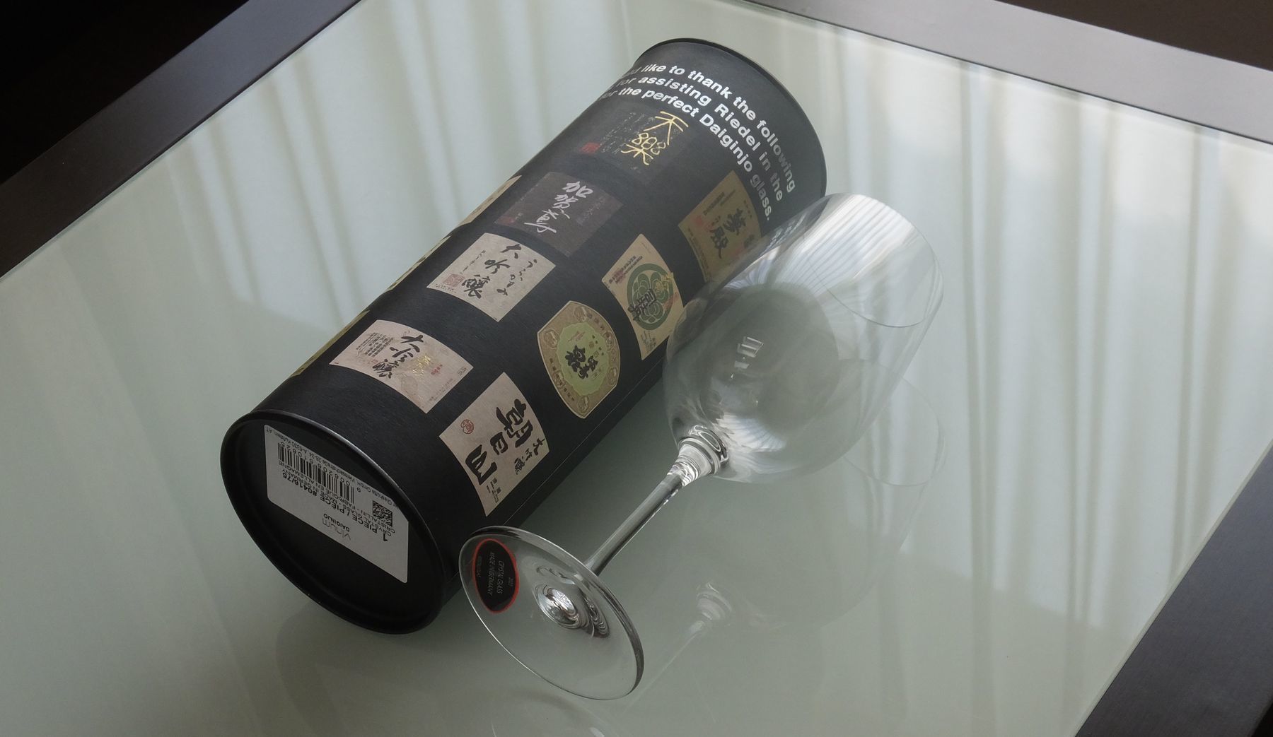 テーブルの上に置かれたリーデル（RIEDEL）『ヴィノム・シリーズ』の大吟醸グラスと、専用のロゴ入りチューブ缶（筒型ケース）の写真。大吟醸酒の香りを際立たせる卵型の美しいフォルムと、ギフトとしての特別感を高めるスタイリッシュなパッケージが並び、機能性と贈答品としての品格を兼ね備えた日本酒愛好家のための酒器セットのイメージ。