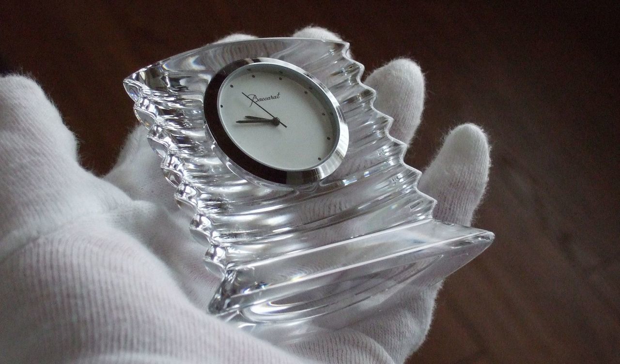 バカラの置き時計、ラランドクロック（Lalande Clock）のクリスタル製クロックを、女性の手のひらにそっと載せている情景を捉えた写真。アール・デコ調の優美なフォルムと、繊細なダイヤモンドカットが特徴的なクリスタルが、手のひらを通して光を透過させ、特別な輝きを放っている。時計の重厚感とクリスタルの透明度が強調され、時間の尊さと不変の美しさを象徴する愛蔵品の情景。