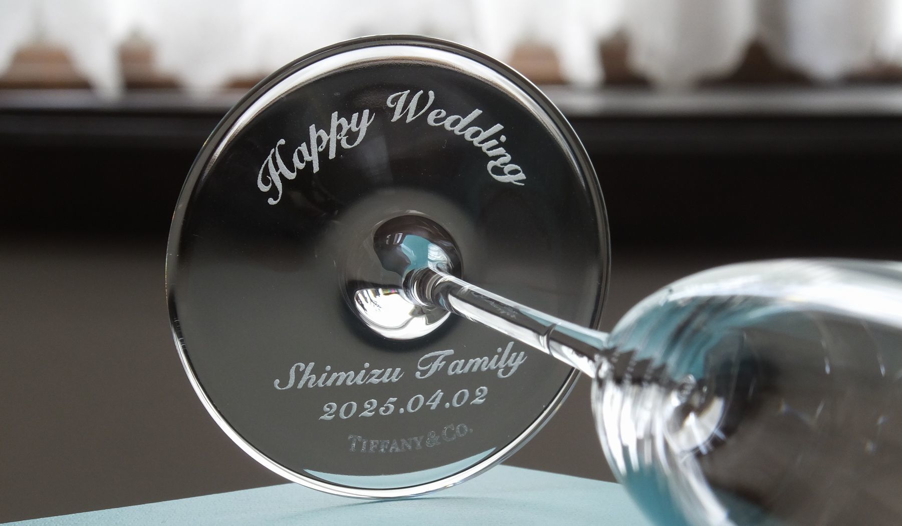 ティファニー（Tiffany & Co.）の最高級クリスタル製シャンパングラスの底面に、『Happy Wedding』のお祝いメッセージ、新郎新婦のお名前、そして大切な誓いの記念日付が精緻に名入れ彫刻された結婚祝いの写真。グラスを傾けるたびに透明感あふれるフット（底面）から愛と祝福の言葉が密やかに覗き、テーブルを華やかに彩るティファニーならではの上品な輝きと、ご結婚のお祝いにふさわしい特別感あふれるメモリアルギフトイメージ。
