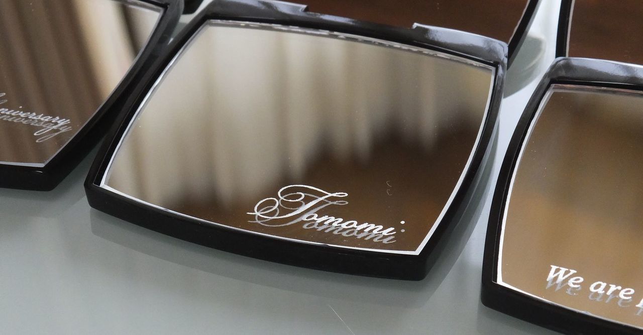 シャネルのコンパクトミラーを開いた際、下部ミラーに『Tomomi』と名入れ彫刻を施した写真。黒を基調としたブランドのデザインに、シルバーに輝く名前の刻印が際立つ、上品で特別な一点物のイメージ。