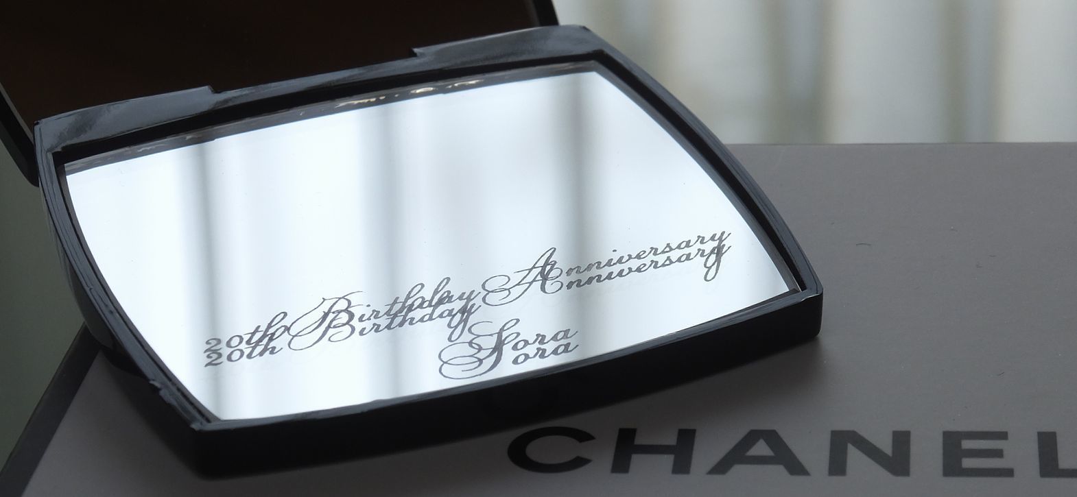 シャネル（CHANEL）のコンパクトミラーのクローズアップ写真。20歳の誕生日プレゼントとして贈られた、黒く象徴的なケースを開いた鏡面、あるいは裏側に、『20th Birthday Anniversary Sora』という成人を祝う記念メッセージと名前が繊細に名入れ彫刻されているのが鮮明に確認できる。ブランドの高級感と、人生の大きな節目である二十歳の誕生日を祝う祝福の想いが込められたパーソナルでエレガントなカスタムギフト。