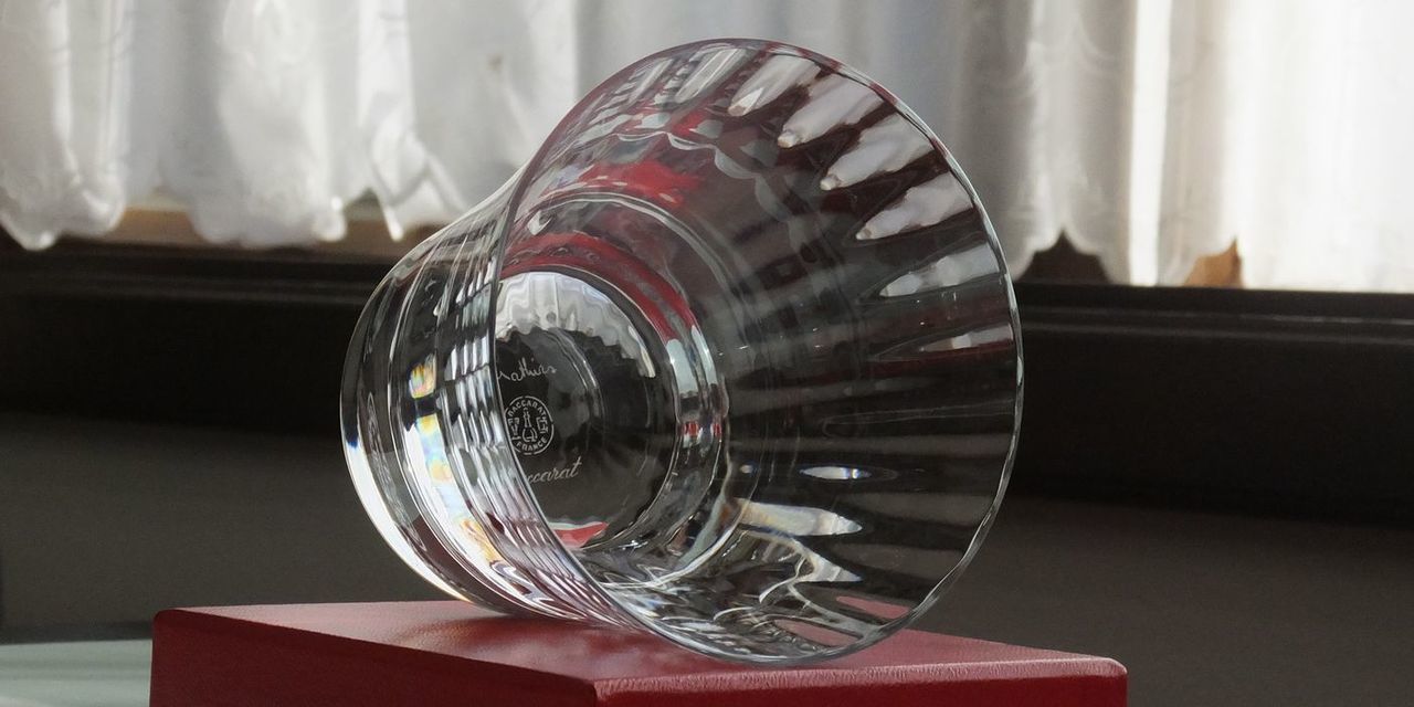 バカラの『ミルニュイ タンブラー』が、バカラのシンボルカラーである赤い純正ボックスの上に、横に倒して置かれている様子の写真。アラビアンナイトをイメージしたミルニュイ特有の優雅なプリーツカットと、深紅のボックスの強烈な色彩のコントラストが際立っている。タンブラーが横向きに置かれることで、そのクリスタルの複雑なカットと輝きがより強調され、最高級ギフトとしての存在感と美しさを視覚的に伝えている。