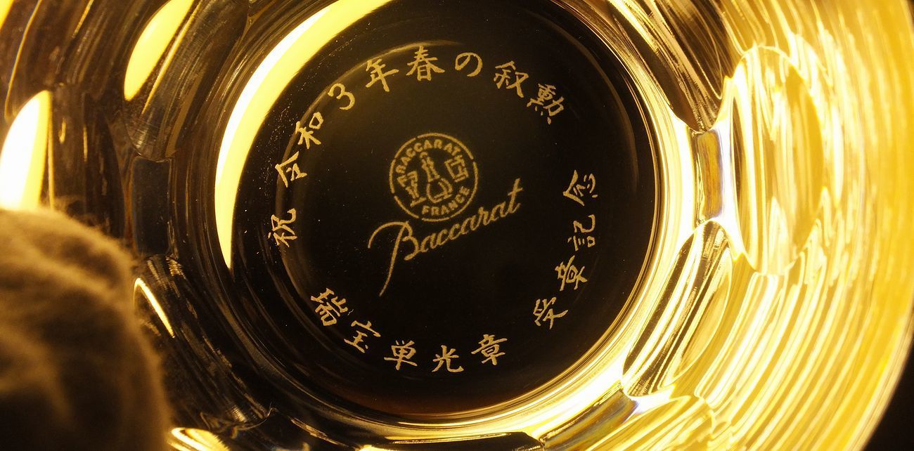 バカラ（Baccarat）の『ベルーガ タンブラー』の底面に、『祝 旭日小綬章受章』の文字と贈り主名が精緻に名入れ彫刻された写真。ラウンドカットの美しいディンプルデザインが光を柔らかく反射する最高級クリスタルグラスの底から、叙勲・褒章の栄誉と祝福のメッセージが密やかに輝き、一生ものの偉業を讃える最高峰の受章記念品・お祝いギフトにふさわしい格調高いイメージ。