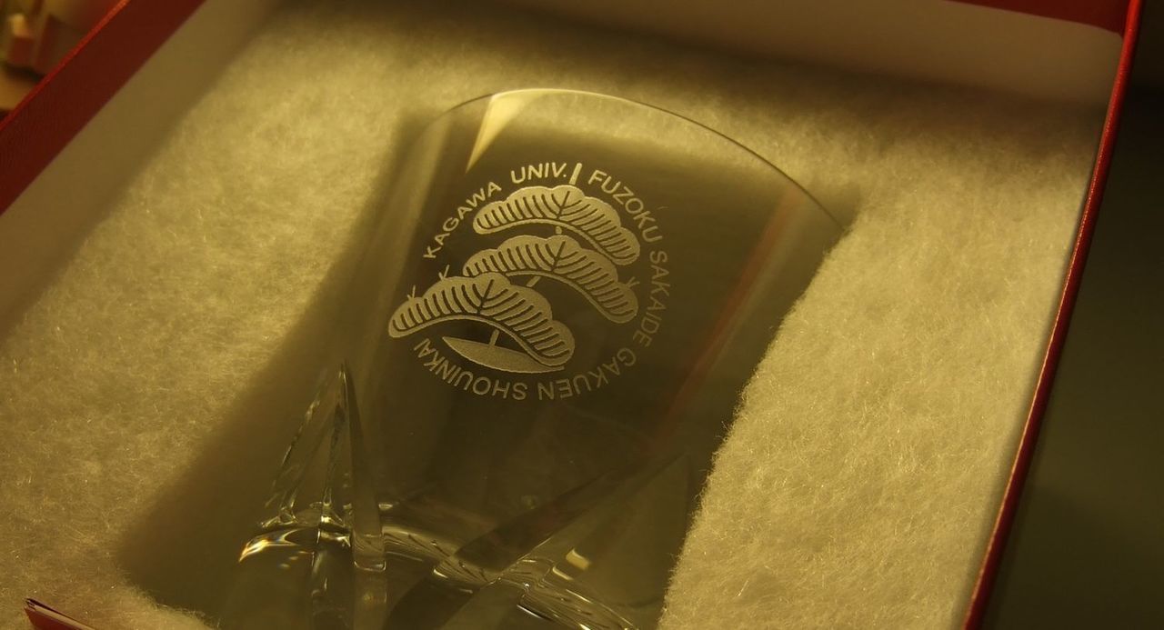 バカラグラスの側面に、精緻なエッチングで校章（ロゴデザイン）と卒業年度、お名前を名入れ彫刻した卒業記念のサンプル写真。クリスタルの高貴な輝きの中に母校の象徴が浮かび上がり、学び舎での思い出と共に歩む新たな門出を祝福する、格調高く一生の宝物となる卒業・周年記念ギフトのイメージ。