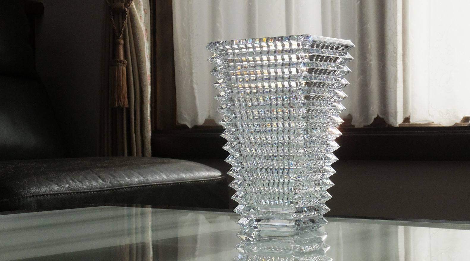 バカラの『アイ ベース スクウェア（角型）』が、モダンなガラステーブルの上に置かれている様子の写真。レンズのカットをイメージした、内外に施された凹凸が、光の屈折により万華鏡のような視覚効果を生み出している。透明なクリスタルと透明なガラステーブルが組み合わさることで、ベースの幾何学的なカットの美しさと複雑な光の反射が最大限に強調され、空間にアートな存在感を与える洗練された雰囲気を醸し出している。