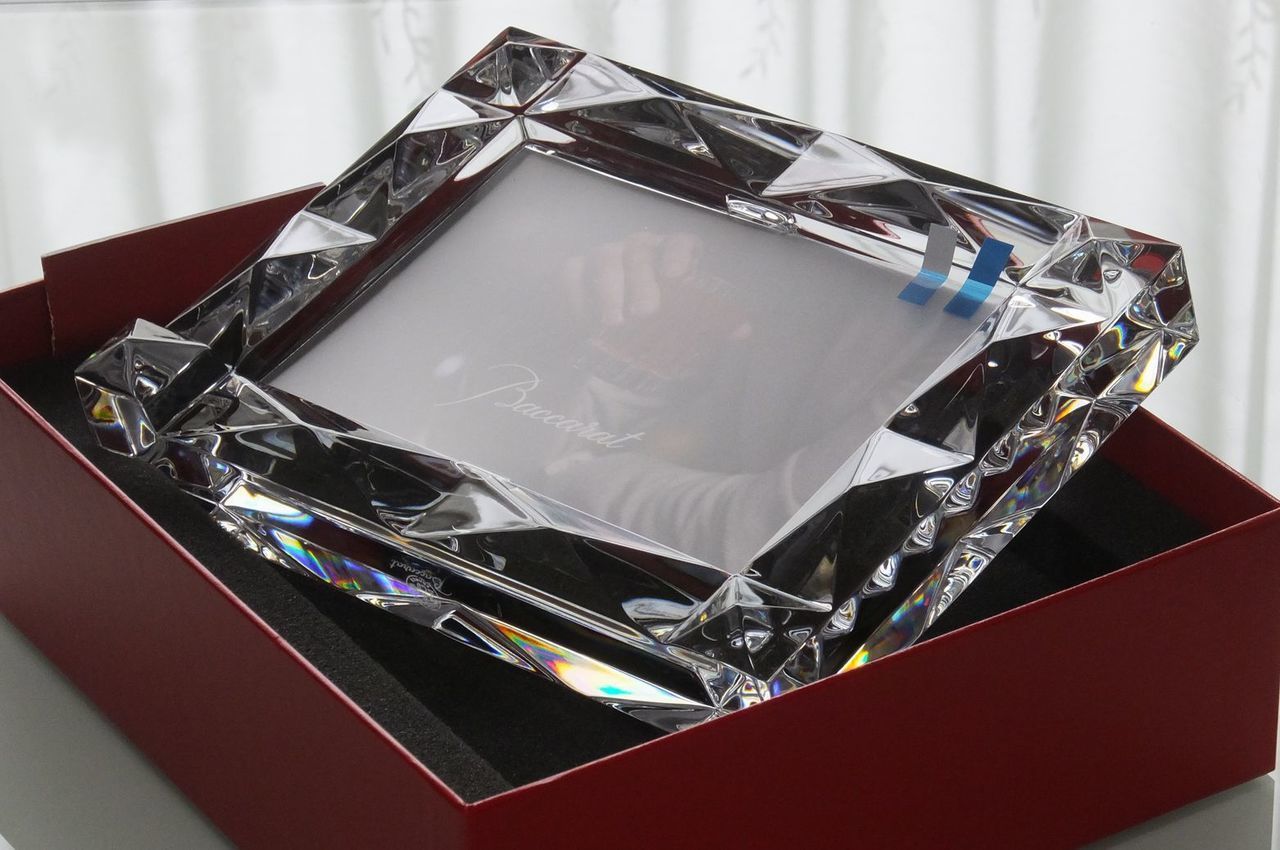 バカラ（Baccarat）の象徴的な赤いギフトボックス（レッドボックス）に収められた最高級クリスタル製『ルクソール フォトフレーム』の写真。古代エジプトの建築から着想を得たピラミッド状の立体的なダイヤモンドカットが光を放ち、ブランドを象徴する深紅のボックスがそのラグジュアリーな存在感を一層引き立てる、大切な記念日の贈り物や格調高い名入れギフトに最適な最高峰のインテリア・贈答品イメージ。