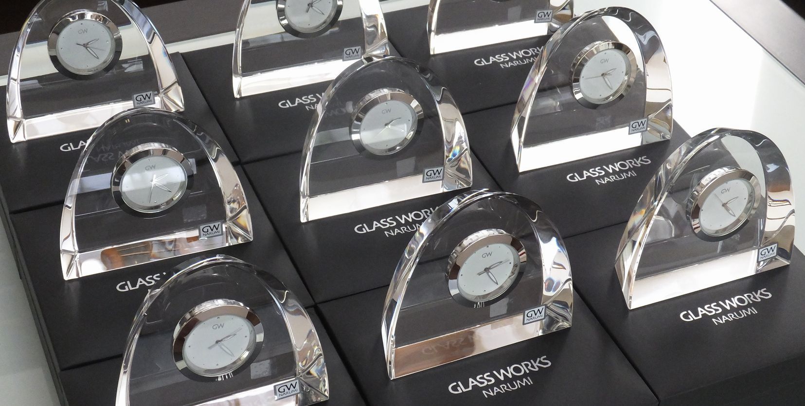 テーブルの上に、グラスワークスナルミのアーチクロックが大量に整然と並べられている写真。透明感のある光学クリスタルの時計が光を反射して輝き、企業の創立記念や永年勤続表彰などのために多数用意された記念品の壮観な様子を伝えるイメージ。