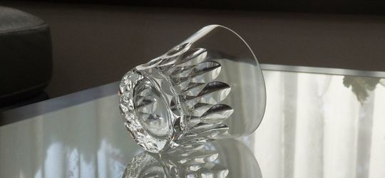 バカラのイヤーグラス、ダリア（Dahlia）タンブラー2026のクリスタルグラスが、テーブルの上に横に倒して置かれている写真。ダリアの花弁をモチーフとしたダイナミックかつ複雑なV字カットが、長い側面を通して光を反射させ、グラスの優美なフォルムを際立たせている。重厚な底面と、繊細なカットが描く光の軌跡が、クリスタルガラスの最高峰としての存在感と芸術的な美しさを強調した情景。