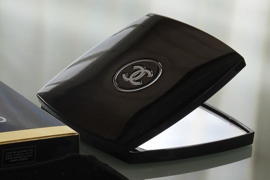シャネルのコンパクトミラーが優雅に少し開かれ、テーブルの上に置かれた状態。高級感漂うデザインが光を反射し、洗練されたエレガンスを感じさせる