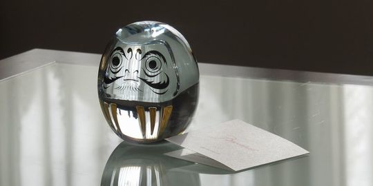 バカラのクリスタル製のダルマがテーブルの上に置いてある写真。透明なクリスタルで象られたダルマが、光を反射して輝き、日本の伝統的な縁起物とフランスのクリスタル技術が融合したユニークな魅力を放っています。名入れギフトとしても、特別な願いを込めるお守りとしても最適な贈り物です。