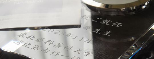 グラスワークスナルミのエンバシークロックに、『教授ご就任』の祝辞、先生のお名前、『2025年11月 東北医科薬科大学 消化器内科一同』と名入れ彫刻を施した写真。透明度の高い光学クリスタルに、医局員一同からの敬意と祝意が刻まれた、就任祝いに相応しい格調高い記念品のイメージ。