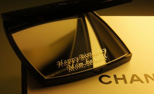 シャネルの名入れコンパクトミラーが少し開いた状態で、鏡面には『Happy Birthday Mom, be well』の彫刻メッセージ入り。大切なお母さんへの誕生日プレゼントにふさわしい、上品で特別感のあるオーダーメイドギフト。