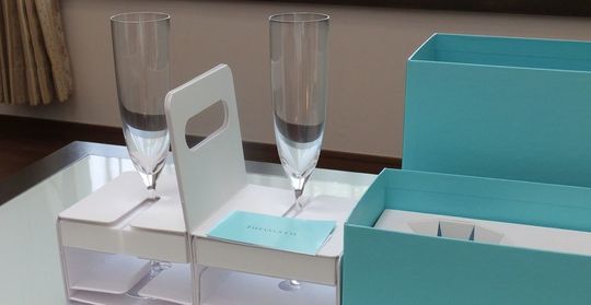 ティファニー商品一覧ページ：ティファニーホームエッセンシャルシャンパンフルートペアを、ティファニーブルーボックスから取り出した瞬間の写真。ペアグラスが梱包材にしっかりと収まっている様子が分かり、丁寧な梱包とギフトの準備が伝わります。