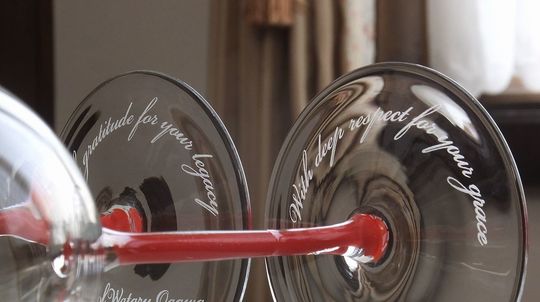 リーデルの赤い脚が特徴的なワイングラス2脚に、それぞれ「gratitude for your legacy」と名前、もう片方には「With deep respect for your grace」と名前が底面に彫刻された、退職祝いに贈るパーソナルな名入れギフトの写真。