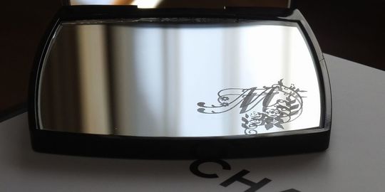 シャネルのコンパクトミラーに、装飾的なデザインで彩られたイニシャル『M』を名入れ彫刻したアップの写真。洗練されたアイテムに、より一層エレガントでパーソナルな魅力が加わっています。