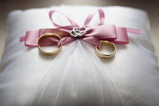 白いリングピローにピンクのリボンで結ばれた、ゴールドの結婚指輪2つ。結婚式やエンゲージメントフォトに最適な、ロマンチックなウェディングリング。