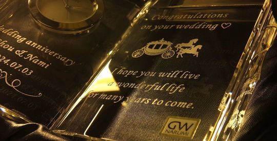 グラスワークスナルミのディクショナリークロックの両面に名入れ彫刻が施された特別な記念品。左面には「Wedding anniversary」の文字とお二人の名前、記念日が彫刻され、右面にはお祝いのメッセージと馬車のデザインが刻まれた美しいギフト