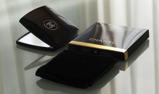 シャネルのコンパクトミラーの一式紹介写真：箱、ミラー本体、そしてミラーを収納するベロア風の袋が、テーブルの上に美しく配置された写真。ギフトとしての完全なセットと、ブランドの細部へのこだわりを伝えます。