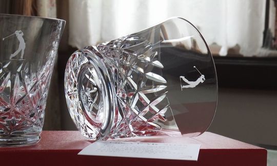 バカラのアリア（Aria）タンブラーのクリスタルグラスのクローズアップ写真。グラスの側面に、ゴルフ記念品ギフトとして『ゴルファーのスイングをしているイラスト』が名入れ彫刻されているのが鮮明に確認できる。しずくを思わせる繊細なカットが特徴のアリアタンブラーのなめらかな面に、躍動感あふれるイラストと『2025』の文字が刻まれ、ゴルフコンペの優勝や特別なホールインワンを記念する世界最高峰の輝きを持つ記念品。