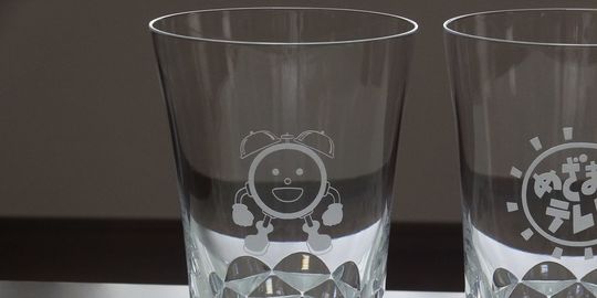 バカラのハイボールグラス、スウィングハイボールの側面に、めざまし君のキャラクターデザインとめざましテレビのロゴが名入れ彫刻された写真。グラスのなめらかな曲線と、親しみやすいキャラクターとロゴが融合し、特別な記念品としてのユニークさと、番組への愛着が伝わるデザインとなっています。