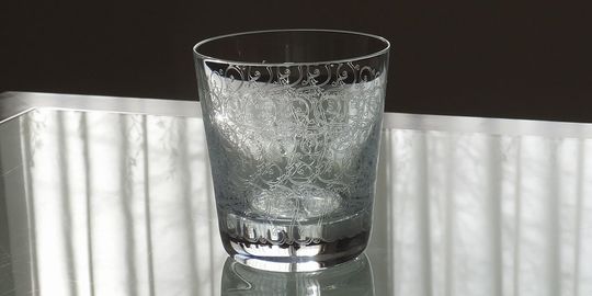 バカラのタンブラー（ロックグラス）の名入れページの商品一覧で使用する写真。バカラ ローハンタンブラーがテーブルの上に置かれています。アシッドエッチングによる繊細なアラベスク模様が特徴的なグラスが光を反射して輝き、名入れギフトとしての優雅さと伝統的な美しさを演出しています。