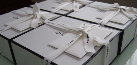 白いシャネルのリボンが飾られた特別仕様ギフトボックスが並べられ、贈り物としての洗練された魅力が伝わるシーン。上品で高級感あふれるシャネルのギフト。